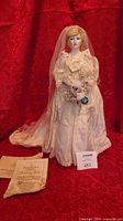 Full view of doll standing on red backdrop wearing gown and veil, certificate visible