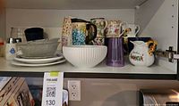 Cabinet shelf showing assorted mugs, bowls, plates, creamer and tumbler