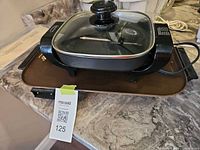Square black electric skillet with glass lid stacked on rectangular brown griddle