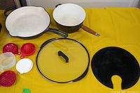 Overview of skillet, saucepan, lid, splatter guard, plastic accessories on yellow surface