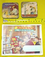 Two square Meccano tins beside ruler and Meccano booklet cover