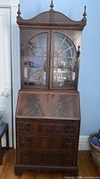 Full view of mahogany secretary desk showing upper glass cabinet, drop-front section, three drawers, finials, bracket feet