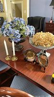 All items on dining table: dark urn with blue hydrangeas, brass candlesticks with candles, silver-plated tray on stand, dried hydrangeas in background