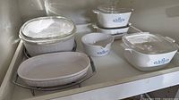 Group of CorningWare pieces on shelf: round casserole with lid, two square casseroles with lids, gravy boat, oval baker, two chrome cradles