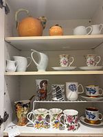 Cabinet view showing pumpkin teapot, sugar bowl, creamers, pitcher, floral rose teacups and various mugs