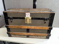 Front view of trunk showing canvas body, wood slats, black metal trim, brass lock