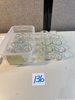Plastic bin containing majority of the small clear glass jars and separate grouping of cylindrical jars