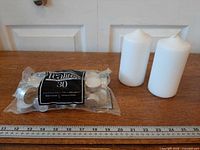 Bagged 30-pack of Tealites tealight candles beside two white pillar candles with ruler for scale