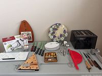 Full lot overview on table showing toaster, apple peeler box, boards, knives, utensils