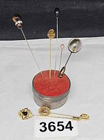 Pins inserted in red pin cushion on metal box with two ornate pins in foreground