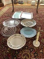 All items laid out on rug: three rectangular trays, two glass spiral platters, three ceramic round platters, towel rack