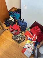 Group of assorted bags on floor including DC backpack, blue briefcase, tapestry bag, teal/purple backpack, reusable totes