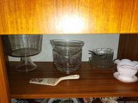 Group of clear glass pedestal compote, pressed bowls, milk glass bowl, ceramic pie server and teaspoons on shelf