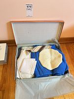 Open box with blue knitwear, cream beret, tie, handkerchief visible