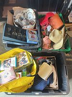 Overview of two totes containing wine glasses box, plates, clock, tin, bag with media, textiles, hat visible