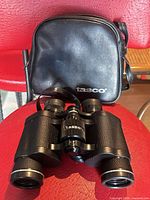 Front view of black Tasco binoculars resting on seat with matching vinyl Tasco zip case behind
