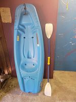 Full view of blue Pelican Solo kayak standing upright next to white/blue paddle