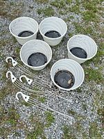 Five white plastic hanging planters with black saucers and five white hangers displayed on gravel