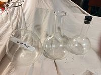 Three round-bottom flasks on white fabric; left flask labeled, right flask with stopper