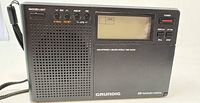 Front view of Grundig G8 radio showing speaker grille, LCD display, power and band buttons, wrist strap