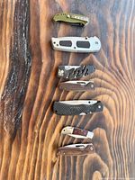 Full view of six folding pocket knives arranged vertically on wood surface