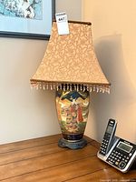 Full view of lamp showing painted ceramic base and tan brocade shade with beaded fringe