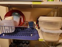 Shelf holding plasticware: Rubbermaid container, pitchers, blue dish drainer with tray, red bowl, blue tumbler, floral plate