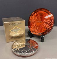 Front view of three cosmetics: Dior compact, L’Occitane boxed cream, Clarins leaf compact