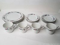 White china set displayed including stacks of dinner plates, salad plates, and saucers with four teacups in front, all with floral pattern and silver rims.