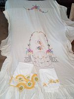 Full view of the vintage bed linens lot laid out, showing the ruffled bed cover with central embroidered girl and floral design, two pillowcases with yellow flower embroidery, two pillowcases with yellow and green tatted trim, and the matching bed runner.