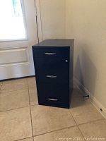 Black metal file cabinet positioned near a door on tiled floor showing front and side view. Three drawers with chrome handles are visible, top drawer lock present without key.