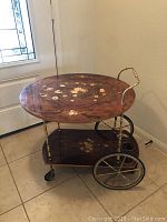 Full view of the inlaid wood tea cart with floral top and bottom shelves, brass handle and wheels