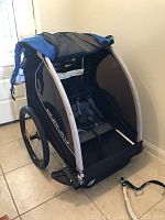 Front and side view showing the trailer with mesh panels, two child seats with harnesses inside, and overall construction and color.