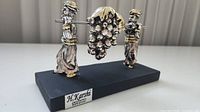 Front view of figurine showing two silver-gilt figures carrying grape cluster on pole, black base with maker plaque