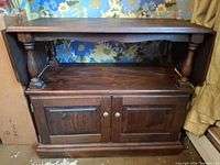 Front view of brown wood drop-leaf serving table showing top shelf, turned supports and cabinet doors