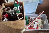 Cardboard box of multiple wrapping paper rolls beside plastic tote of gift bags and accessories