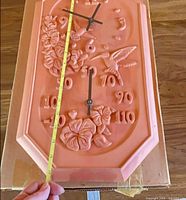 Front view showing clock face, thermometer scale, floral design and tape measure along height