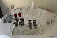 All items laid out on table: tray with silver-rimmed cordials in front, smoked goblets, pitcher with stirrer, red shot glasses, pilsners, wine glasses and clear tumblers visible