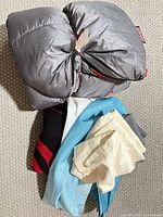 Rolled gray Coleman sleeping bag with red piping, blanket, sheets and curtain grouped on carpet