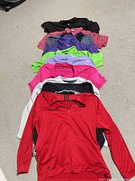 Nine stacked tops showing variety of colors and styles, red Adidas quarter-zip at front