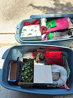 Two totes filled with assorted Christmas wrapping supplies including bags, boxes, ribbons, tissue paper and faux greenery; visible Sundance box