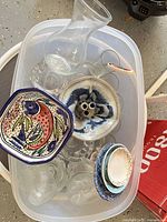Plastic bin containing clear glass vase, glass tumblers, square fish dish, round multi-hole ceramic piece, stack of small ceramic plates/bowls