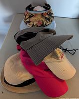 Stacked view of several hats showing grey boonie hat, floral visor, pink, beige and straw hats