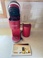 Front view of red Keurig K-Mini Plus with drip tray, pod well open, power cord and manual visible