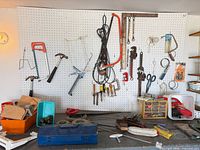 Pegboard wall with hacksaws, hammer, carpenter square, clamps, pipe wrench, screwdrivers visible; bench with toolbox and brushes