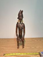 Front view of seated wooden figure showing overall form, headdress, and tooth necklace with scale