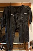 Two black Fox PVC rainsuits hanging, showing full length and overall condition
