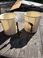 Two brass lamp bases with bulbs and cords alongside two cream fabric drum shades on wooden surface