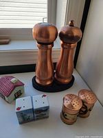 Full lot view showing two tall copper grinders on stand, metal mug-style shakers, ceramic souvenir shakers