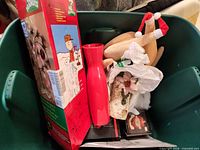 Contents of green tote showing boxed Peanuts tree, red vase, plush turkey with Santa hat, artificial poinsettia, boxed Santa figurine and other holiday items